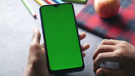 person holding a smartphone with green screen, surrounded by school supplies and an apple.