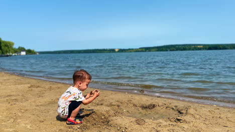little baby boy sits near the river in summer. kid touches the sand, looks at his hands and stands up rejoicing.