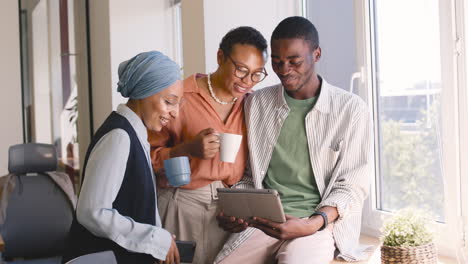 muslim businesswoman, businesswoman and young worker are laughing while looking something at tablet in the office