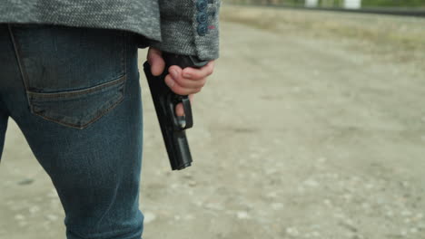 close view of a person in blue jeans and a gray jacket holding a handgun while walking on a dirt road