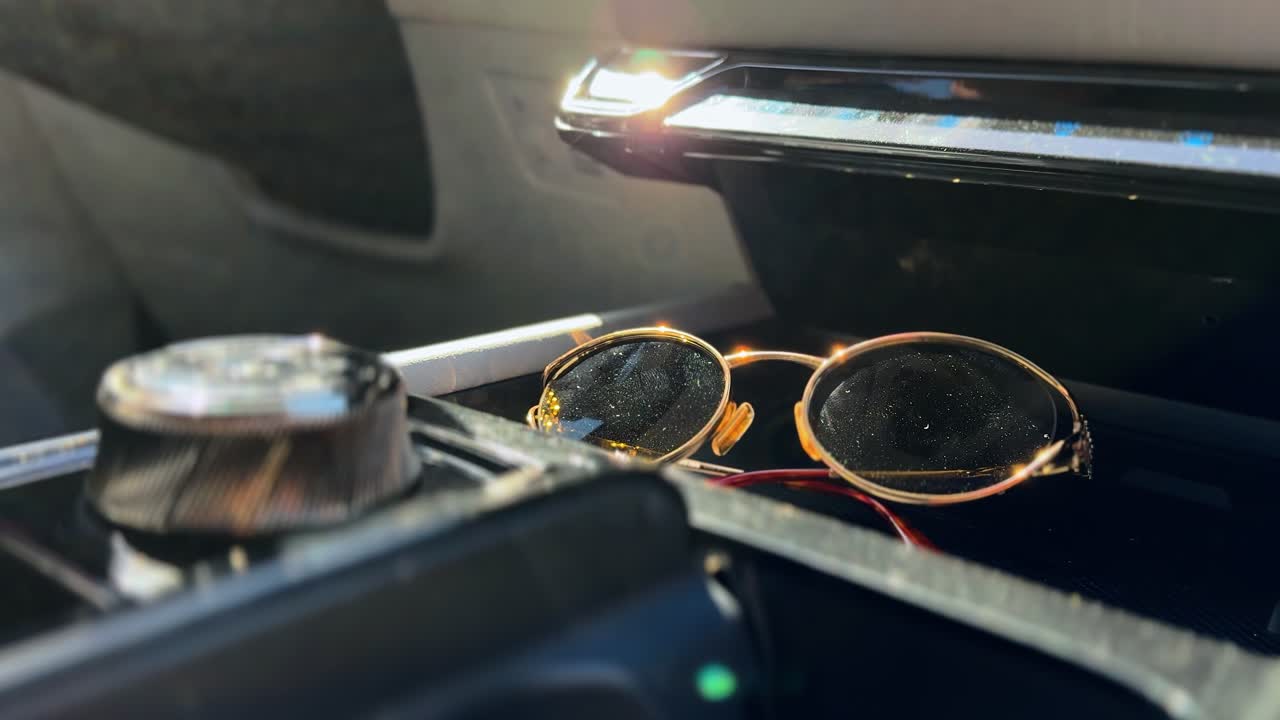 Close-up of a luxury car interior featuring stylish sunglasses and personal travel accessories placed in the center console, illuminated by warm sunlight through the window