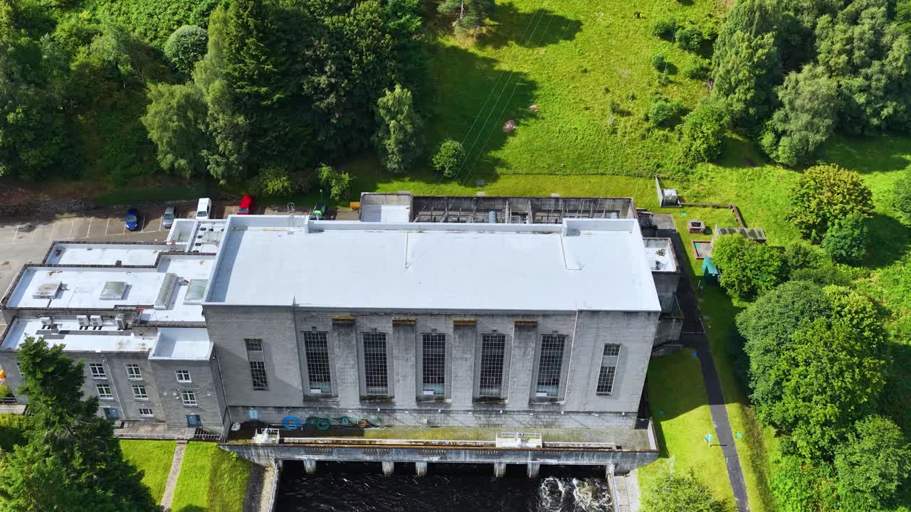 Drone slowly moves above hydroelectric power station, bright daylight, surrounded by lush green landscape