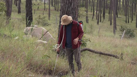 wide long shot of a man working with a backpack sprayer in the forest, he wears a red shirt and hat