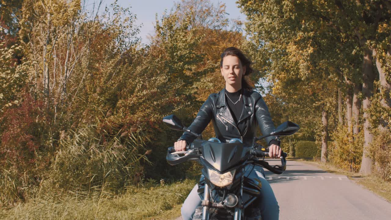 mujer joven europea muy sonriente conduciendo una moto con chaqueta de cuero en el bosque con hojas de otoño doradas vibrantes y coloridas en el día soleado-3