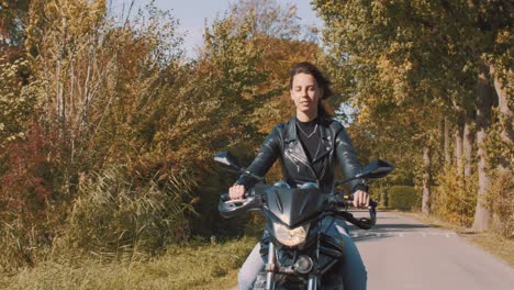 Hübsche-Lächelnde-Europäische-Junge-Frau,-Die-An-Einem-Sonnigen-Tag-Ein-Motorrad-Mit-Lederjacke-Im-Wald-Mit-Lebhaften,-Bunten-Goldenen-Herbstblättern-Fährt-3