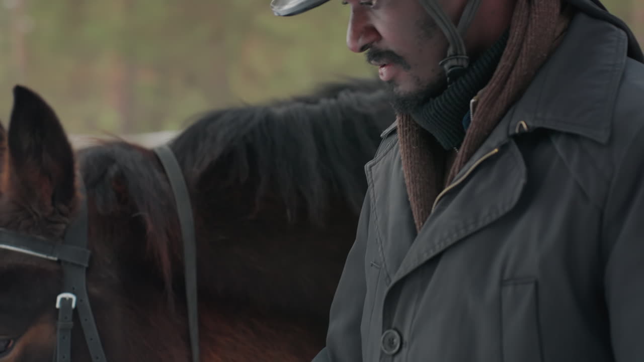 눈 덮인 숲에서 말 기어를 조절하는 남자, 평화로운 겨울 마구간 일상에서 고삐를 점검하는 남성 기수, 겨울 풍경 속 눈 덮인 소나무 숲 한가운데서 말 장비를 주의 깊게 살펴보는 기수