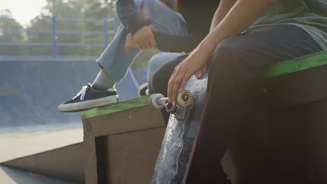 Young-Man-Sitting-With-A-Friend,-Spinning-A-Skateboard-Wheel-In-Skate-Park