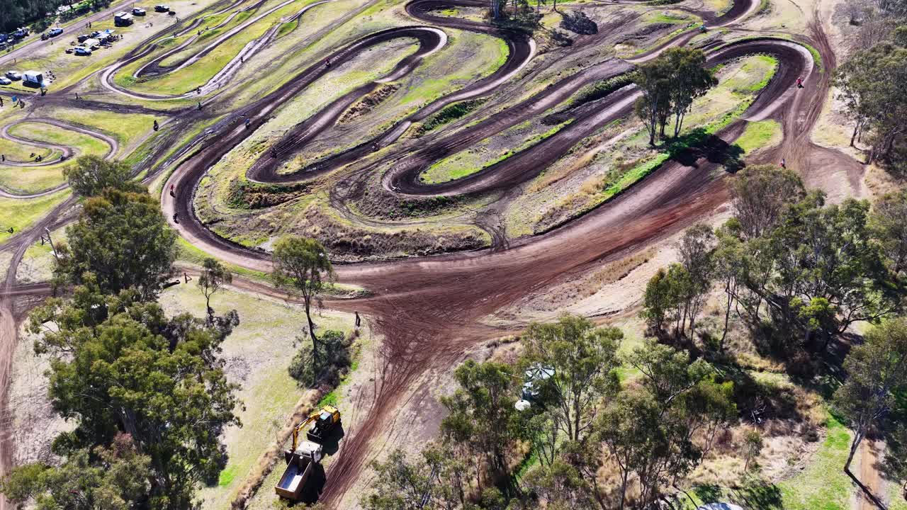 Drone glides above winding motocross track, trees, and parked vehicles under bright midday sunlight