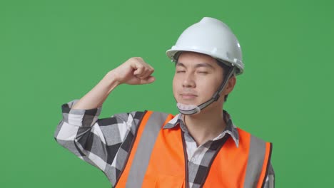 close up of asian male engineer with safety helmet flexing his bicep and smiling to camera while standing in the green screen background studio
