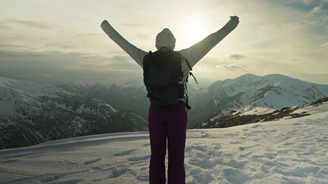 Tracking shot of woman on top of snowy mountain putting her hands up with sun behind, handheld shot
