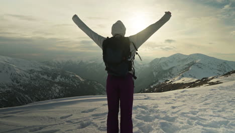 tracking shot of woman on top of snowy mountain putting her hands up with sun behind, handheld shot