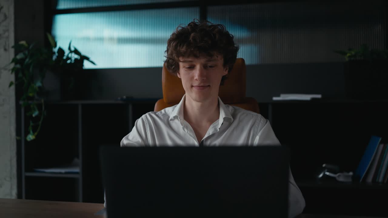 Portrait confident young guy with curly hair in a white shirt posing and working at a gray laptop in a modern office