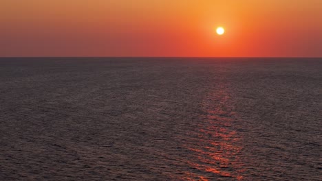 aerial trucking pan above empty ocean with red ball of sun setting with orange gradient in sky