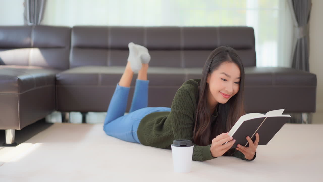Medium close-up young, attractive Asian woman with a hot drink by her side on the floor lies on her stomach as she reads a book