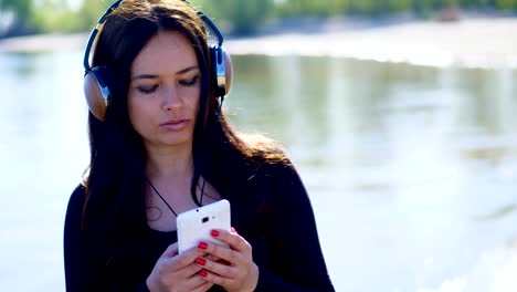 summer, near river, on beach. at sunrise, beautiful woman, with long dark hair, listening to music on headphones from a mobile phone, device, outdoors, dances. enjoying music