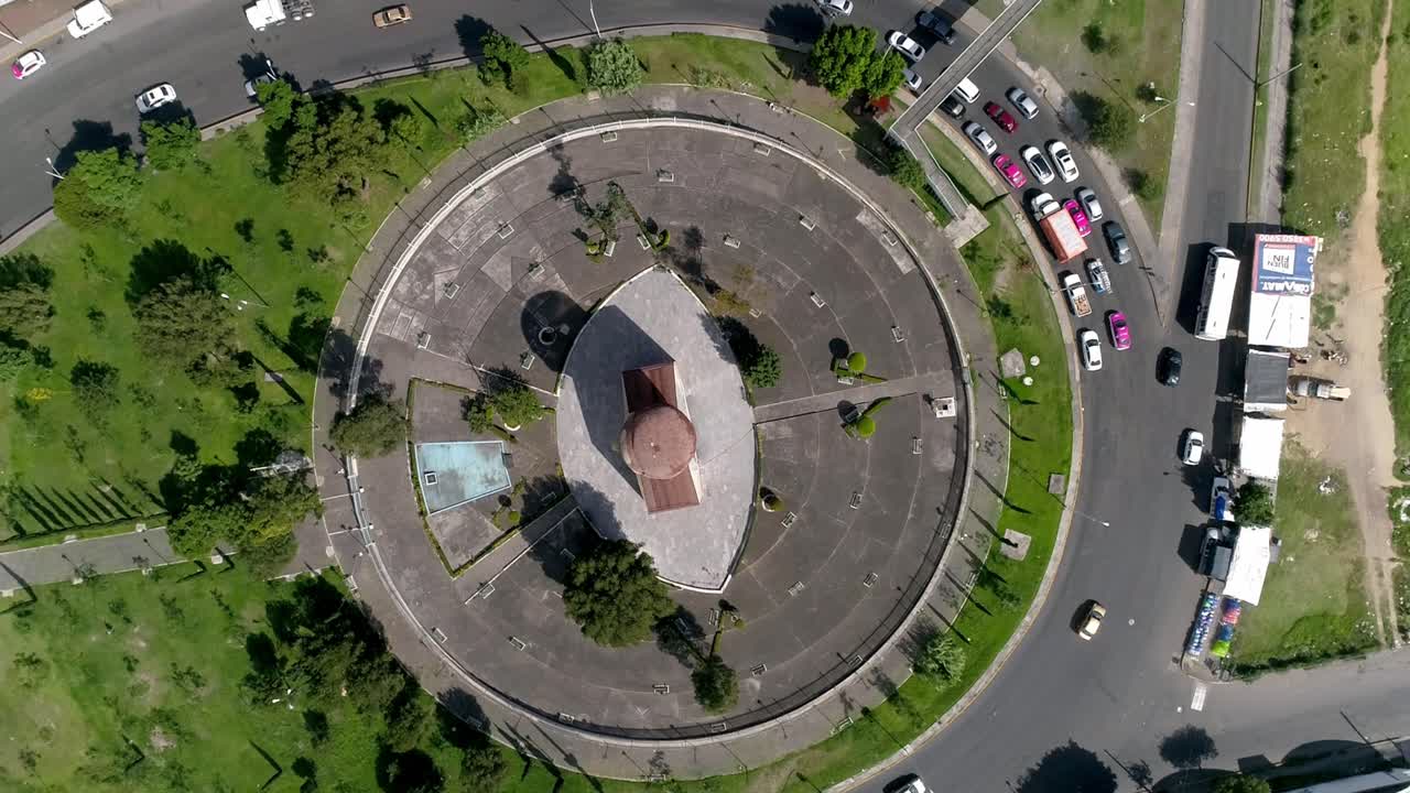 Ripresa aerea con drone di una rotonda con monumento centrale, veicoli che si muovono lungo strade circolari, aree verdi circostanti che mostrano un flusso di traffico organizzato e la disposizione della pianificazione urbana.