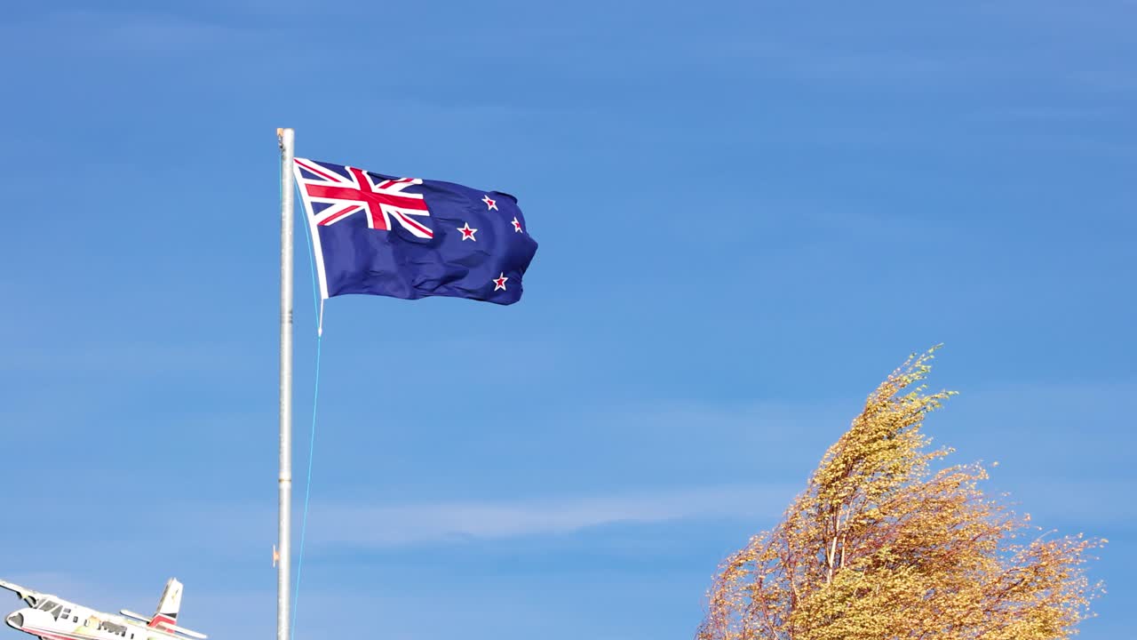 A New Zealand flag waves vigorously atop a tall flagpole against a clear blue sky, with windblown trees and part of an airplane visible, shot in bright daylight
