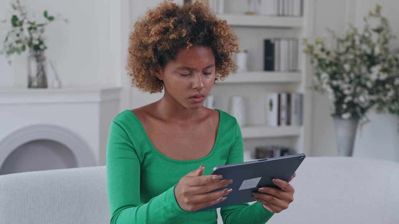 Young dissatisfied african american woman using tablet computer sits on sofa