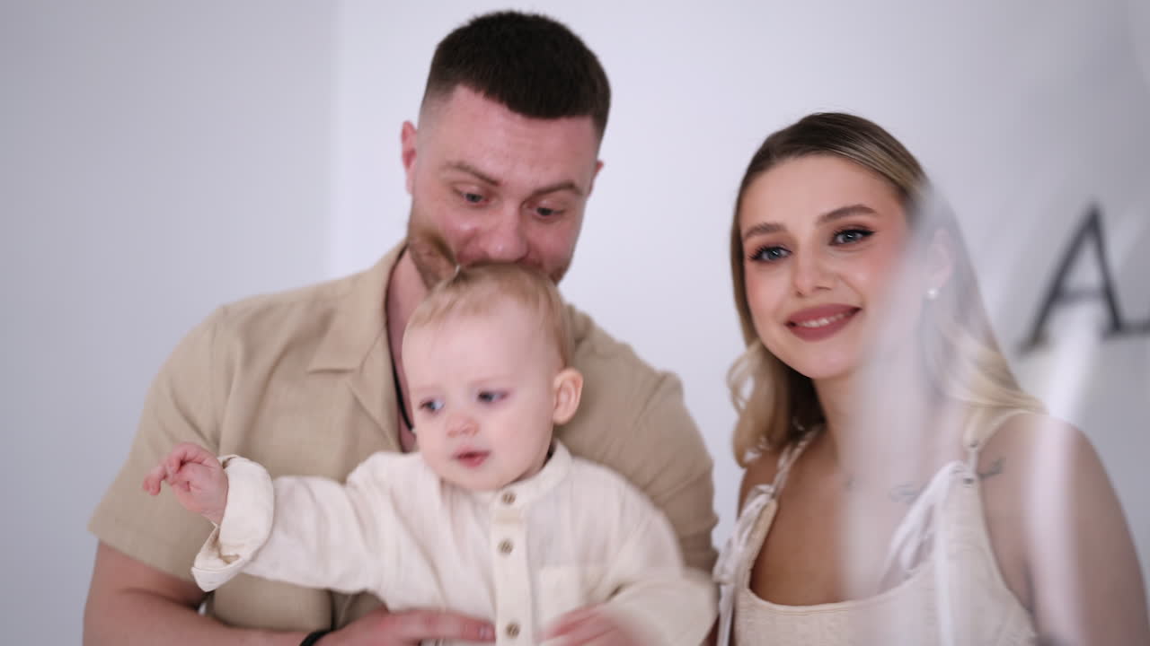 Young Caucasian parents with a beloved baby in hands. Happy mom and dad with their son in studio. White backdrop.