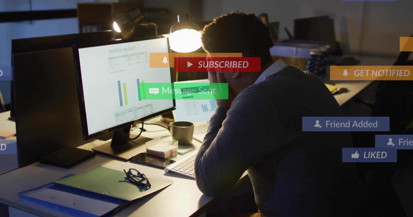 Male worker covering face reviewing monitor charts in office, notifications popping and obscuring