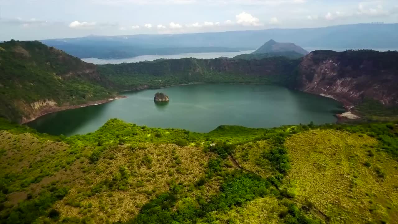 Aerial drone flying over and gliding over Tall lake or Bombon lake in the Philippines during a cloudy day. The lake is green and blue colored and has a giant rock in it while lake is between mountains