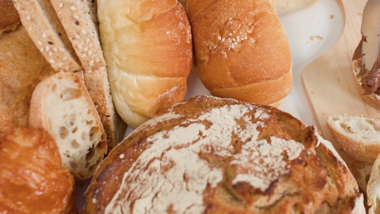 Hands spread chocolate hazelnut cream on bread slices amidst various fresh bakery pastries and loaves.
