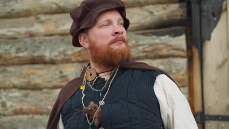 bearded man in medieval clothes stands near wooden house