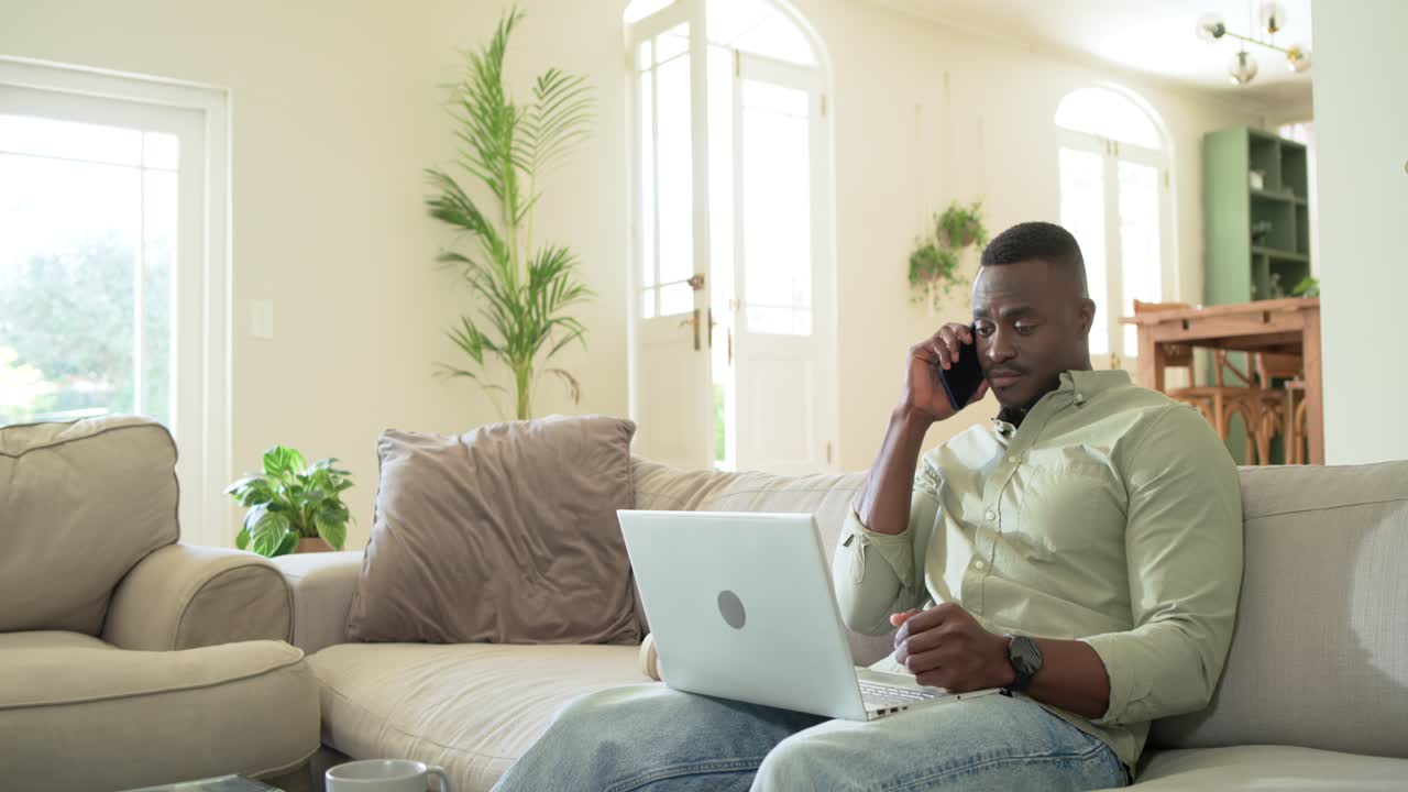Mid adult African American man answering phone checking laptop on couch fixing work issue smiling