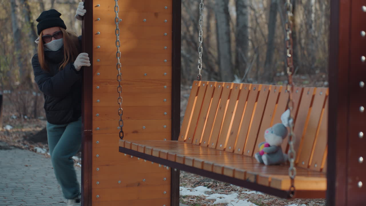 Vaaleaihoinen nainen nappaa nallen penkiltä, puiston keinupenkillä, jossa on ketjut ja puiset säleet. Reppu olkapäillä, käsineelliset kädet tarttuvat pehmoleluun ja nostavat sen kohti taskua. Salakavala vilkaisu ja kiireinen liike.