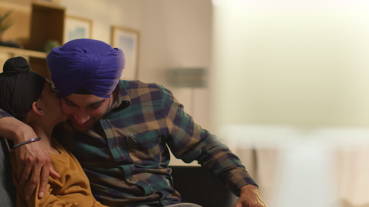 Padre e figlio Sikh affettuosi che indossano turbanti, seduti sul divano a casa mentre parlano insieme.