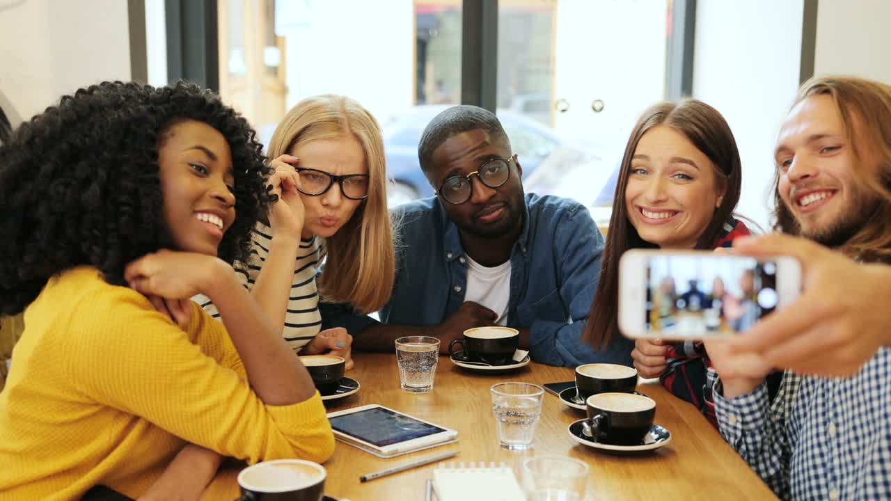 vista de cerca de un grupo multiétnico de amigos haciendo un selfie con un smartphone y haciendo gestos divertidos sentados en una mesa en un café