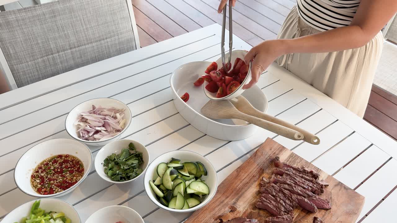 A person in a striped shirt uses tongs to transfer sliced tomatoes into a mixing bowl, surrounded by fresh vegetables, grilled beef, and herbs on a sunlit outdoor table