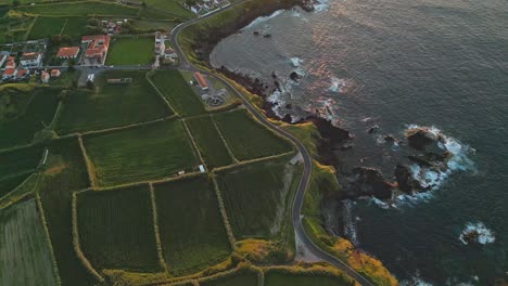 A-coastal-landscape-with-winding-roads,-lush-fields,-and-ocean-waves-at-sunset,-aerial-view