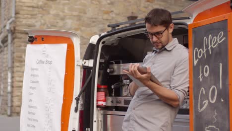 male barista checking tablet leaned against mobile coffee van parked in downtown street,