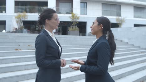 side view of happy female business partners meeting outside, shaking hands, smiling and talking together