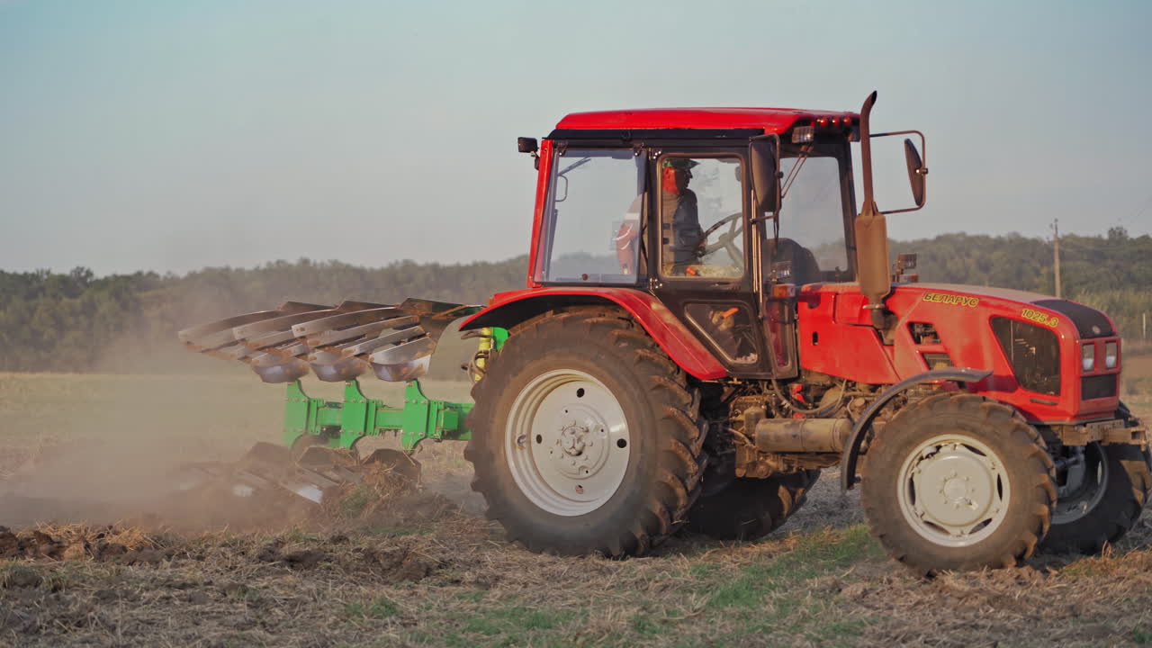 Side view of a red tractor cultivating the soil on the field. Farmer in tractor prepares land for sowing in farmlands outdoors.