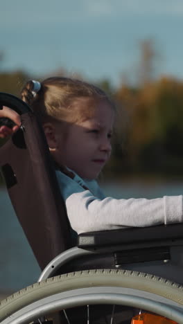 cute preschooler girl with disability looks at flow of water in river on sunny day. child rests with mother in nature on blurred background closeup side view