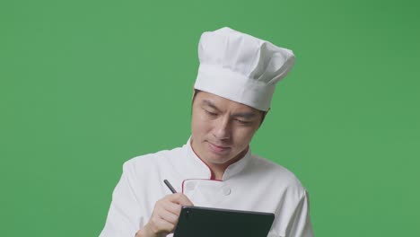 Close Up Of Asian Man Chef Taking Note On A Tablet While Standing In The Green Screen Background Studio