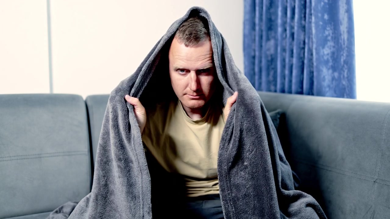 Mand sidder på en sofa, pakket ind i et tæppe, ser seriøs og fokuseret ud, indendørs i en stue om dagen.