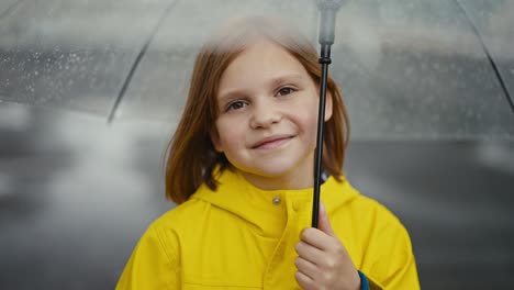 Portrait-of-a-happy-blonde-teenage-girl-in-a-yellow-jacket-holding-a-transparent-umbrella-on-a-walk-after-the-rain