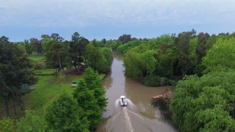 a white boat speeds through a narrow river channel in the paraná delta, flanked by green trees and muddy water under a bright sky, leaving a curved wake, aerial drone follow shot