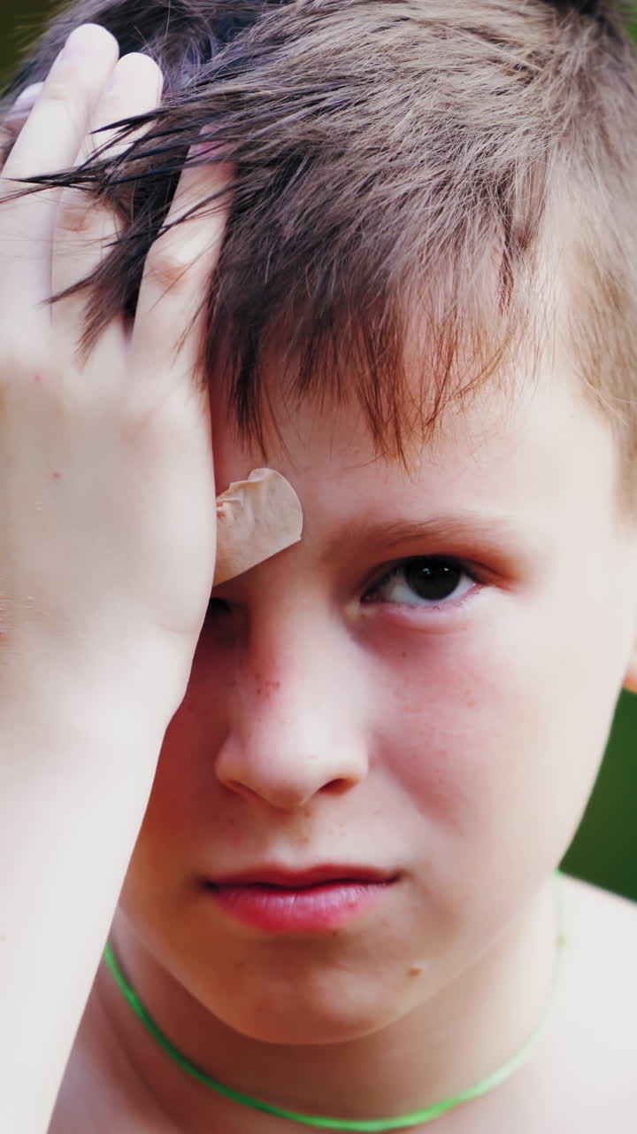 Unhappy boy with closed wound on face outdoors. Young teenager has a pain from his injury on eyebrow. Sad child touches his painful wound in a summertime. Vertical video
