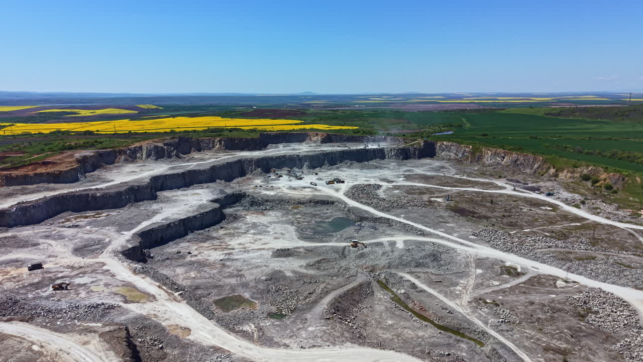 Ampia cava con macchinari e attività di movimento terra, vista dall'alto sotto un cielo azzurro e limpido in una luminosa giornata di sole.