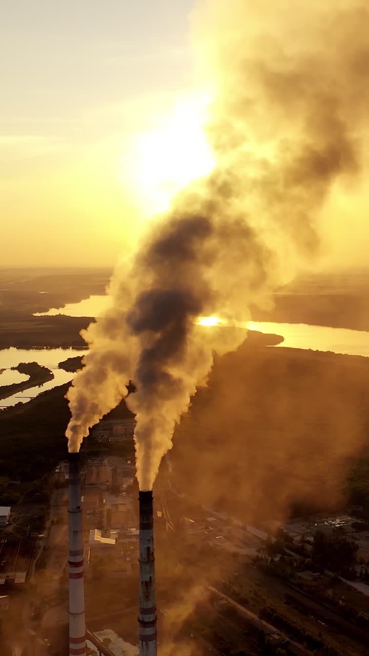 Industrial pipes with smoke in nature. Metallurgical plant among green fields at beautiful sunset. Aerial view. Camera moves right. Vertical video