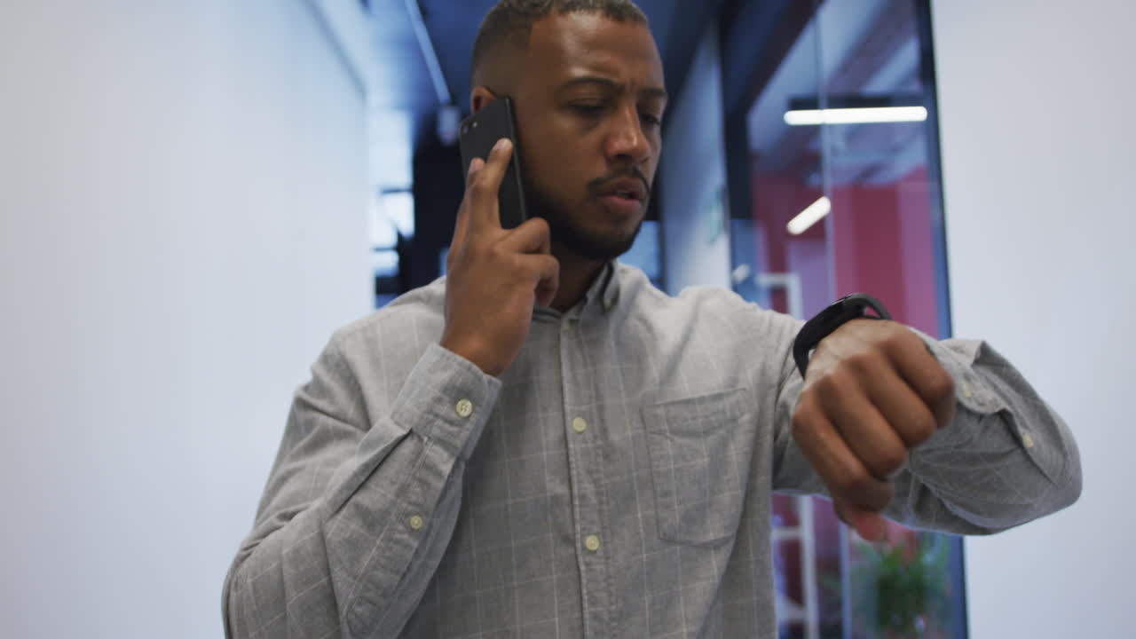 Biracial businessman walking talking on smartphone checking his watch in modern office