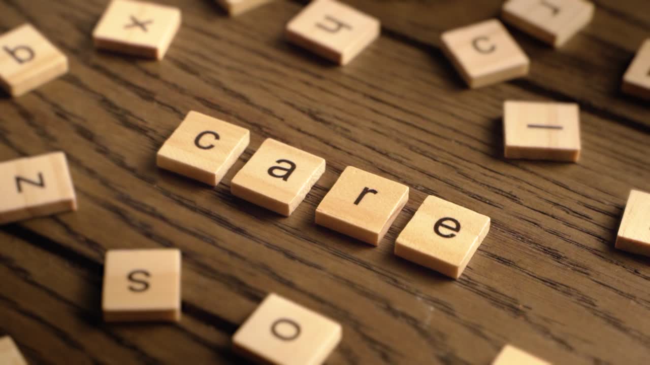 A hand arranges wooden letter tiles to spell the word care on a warm-toned wooden table.