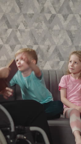 cute toddler gives book to mommy sitting near elder sister on sofa. woman in wheelchair prepares to read interesting story to children before sleep