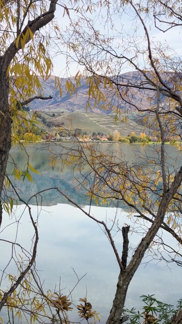 Pemandangan Danau Dunstan yang tenang dibingkai oleh pepohonan musim gugur, menangkap air yang tenang dan perbukitan yang jauh di bawah cahaya matahari yang lembut.