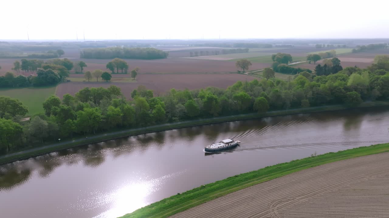 Aerial orbit of a boat cruising a calm river with green farmland in the background.