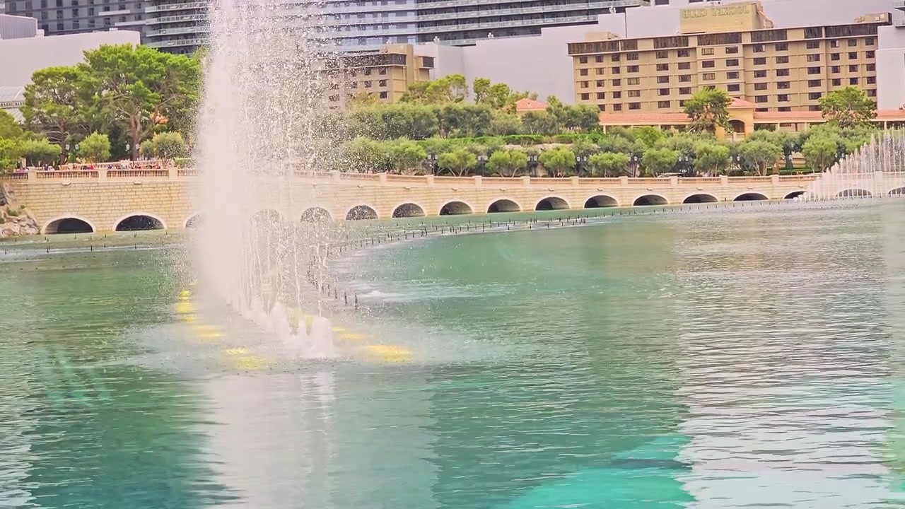 Vista panorámica del Strip de Las Vegas con las Fuentes del Bellagio rociando sobre un lago azul verdoso, con niebla, arcos de piedra, torres de complejos turísticos, árboles, ondas y un ambiente de viaje diurno brillante para el turismo